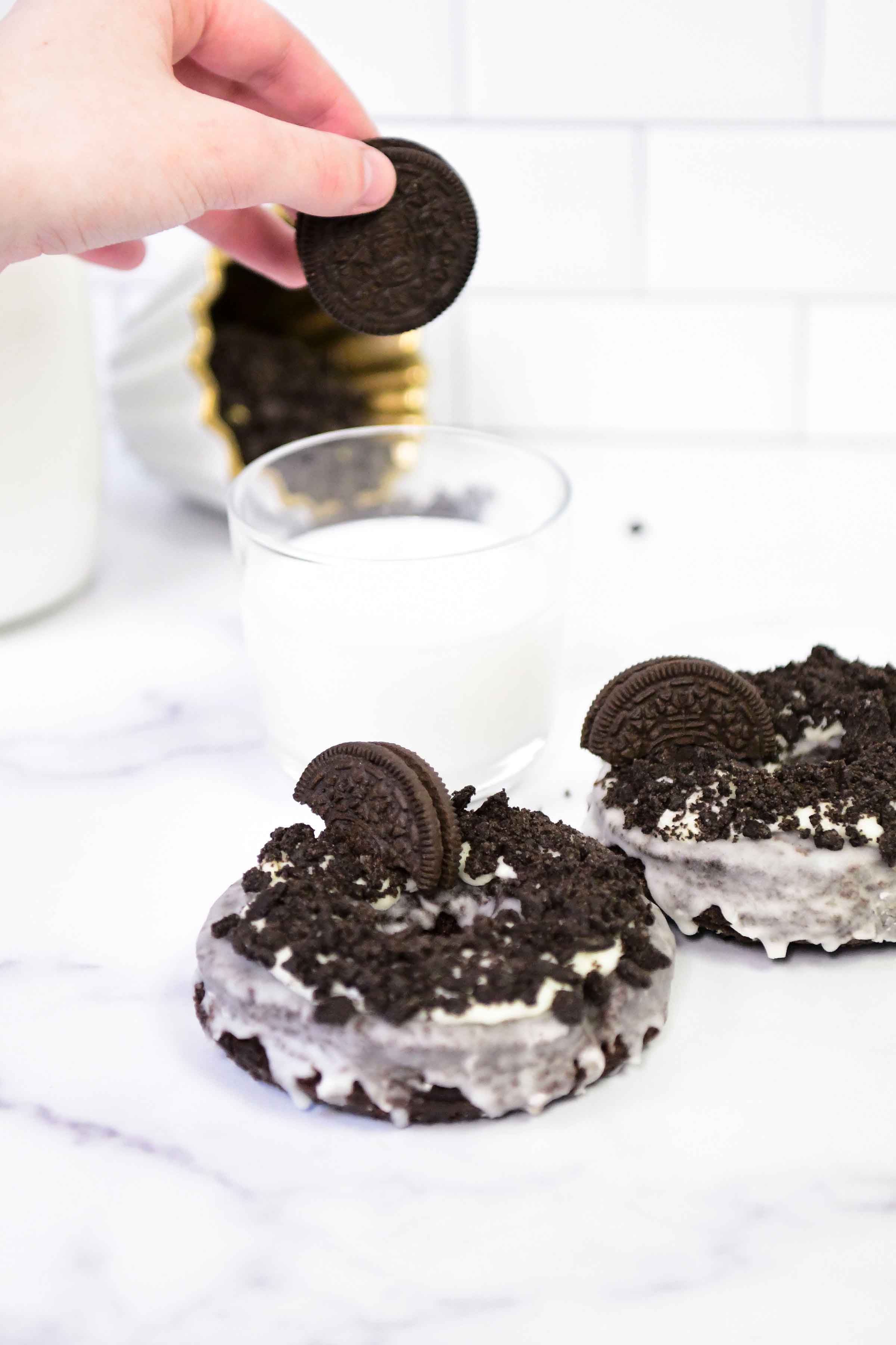 cream and chocolate chips covered donut with an oreo cookie on top and a hand dipping one of the cookies in a milk cup